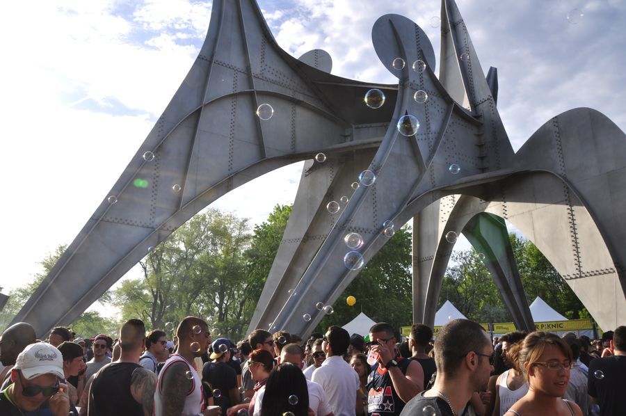 A crowd of people dance underneath a massive metal sculpture with six legs. Above their heads float small bubbles.