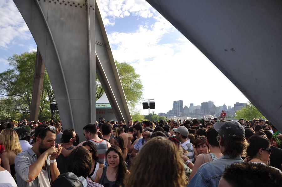 A crowd of people dance underneath a massive metal sculpture beside a large river, surrounded by trees. Across the river, in the distance, are Montréal’s downtown skyscrapers.