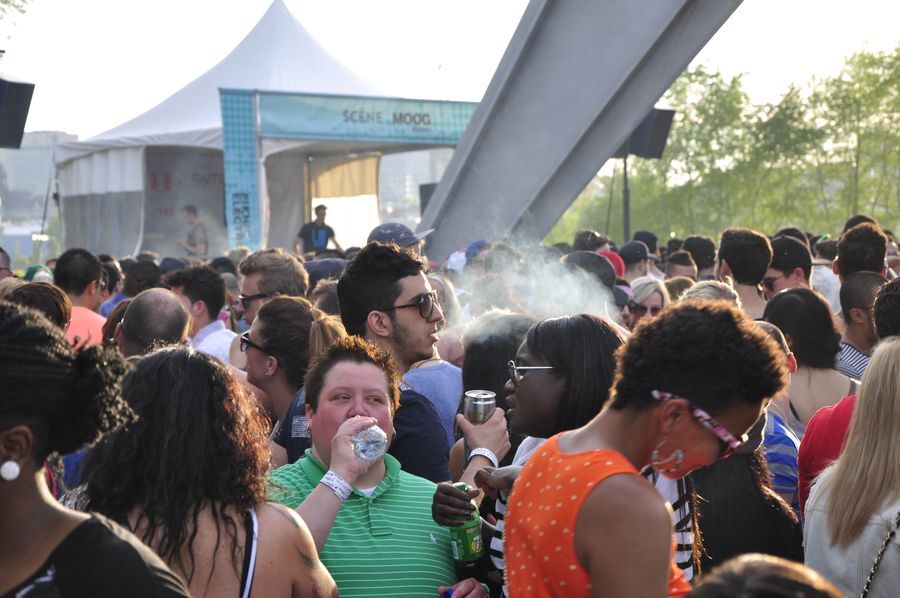 A crowd of people with diverse ethnicities and genders dance underneath a massive metal sculpture. In the background is a raised DJ tent and speakers.