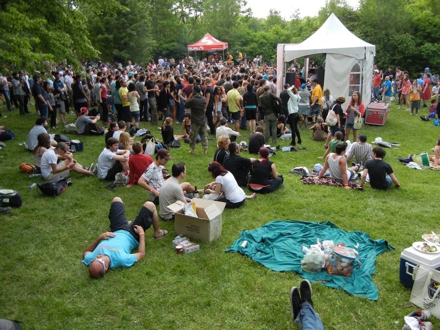 A crowd of people dance around a white tent. The white tent is in the middle of a grassy field surrounded by trees. Some people are laying down on picnic blankets in the field.
