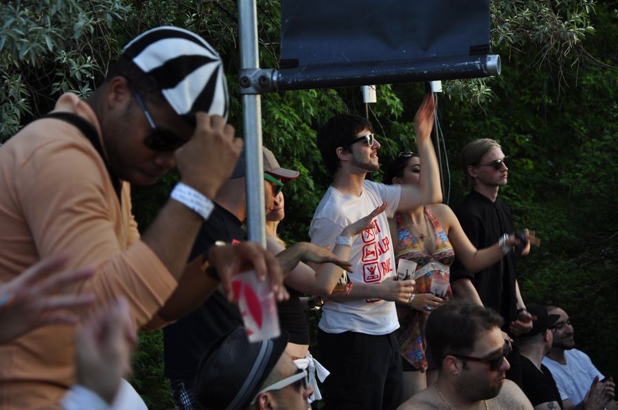 Five dancers face the crowd underneath a speaker. One person of colour wears dark sunglasses and a black-and-white cap. A Caucausian man in sunglasses wears a white t-shirt and raises one hand in the air.