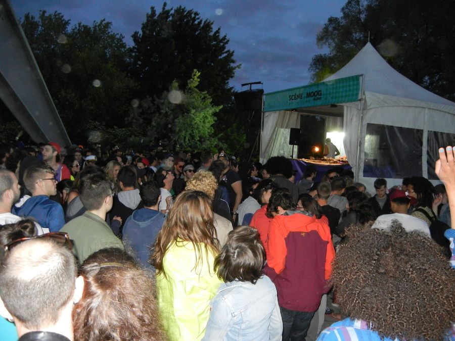 A large crowd of people dance underneath a large metal sculpture at night, facing a raised white tent with a DJ. A banner on the tent reads “Scéne MOOG.”