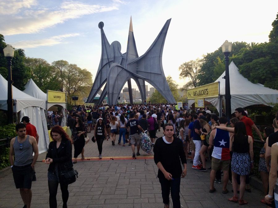 A large metal statue by Alexander Calder towers above hundreds of people on a large paved walkway. On either side of the walkway are white tents selling food and drinks.