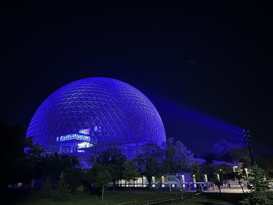 A large geodesic dome is lit by blue lights at night. The dome is surrounded by trees. Inside the dome can be seen a large building with a raised concrete deck.