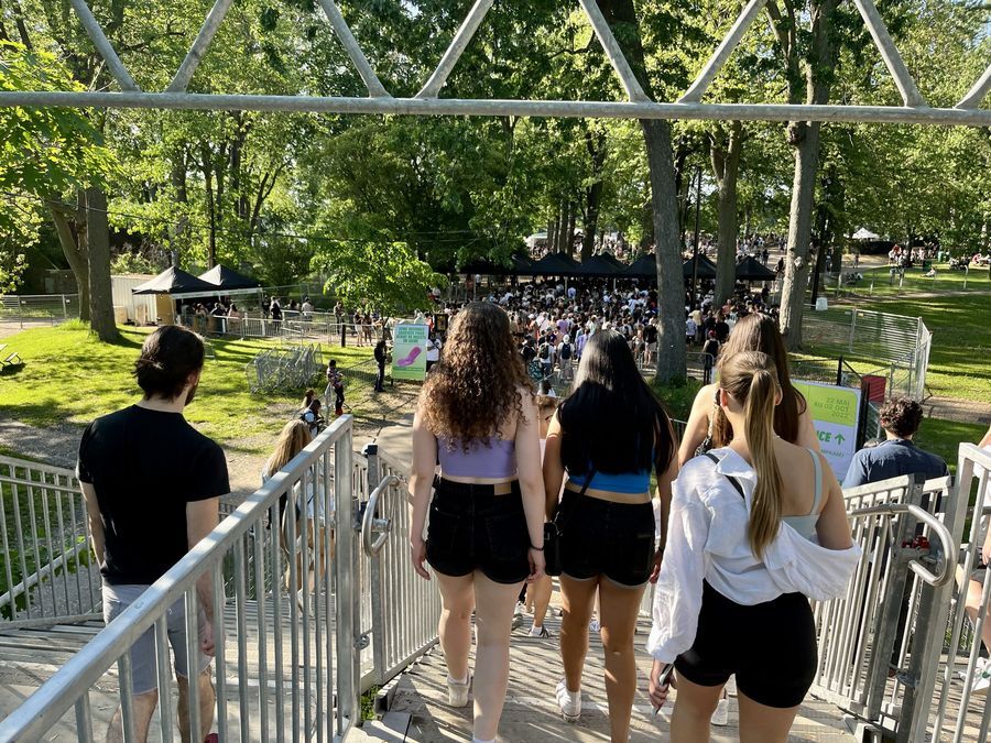 Five people walk down a set of stairs into a grassy area with trees. Four of them are Caucasian women in their twenties and the fifth is a man with dark hair. On the grassy field is wire fencing and several tents.
