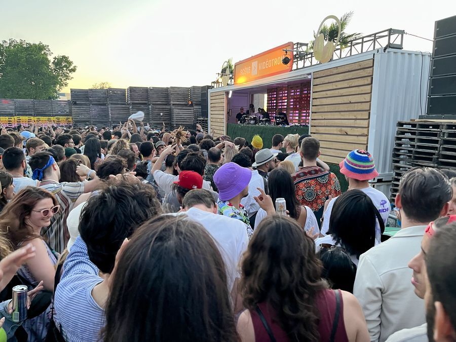 A large dancefloor with several hundred people is surrounded by tall wooden fencing and green trees. A raised stage is flanked by wood panelling and tall speaker stacks. On the stage are the two African American producers known as Octave One and a small crowd of people dancing.