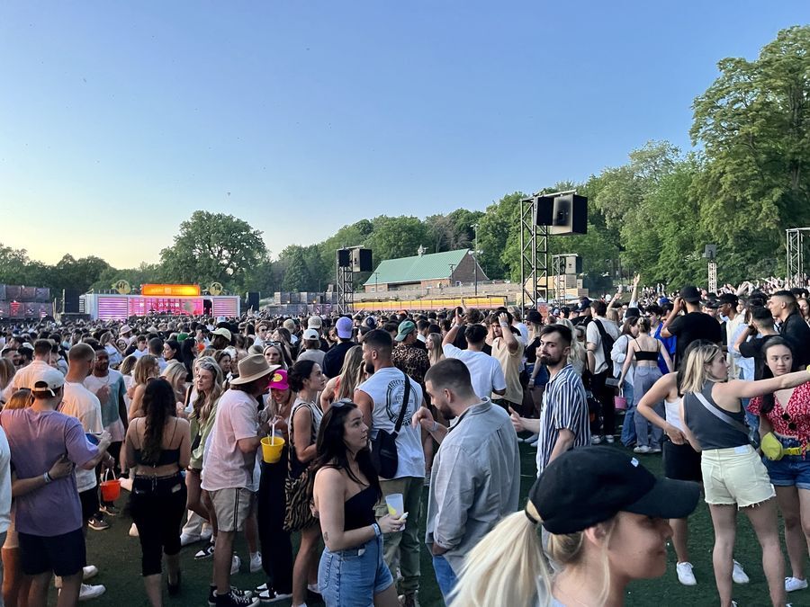 Hundreds of people dance on a large open-air dancefloor surrounded by trees. The DJ booth can be seen in the distance hundreds of feet away. It is lit pink. Speakers hang from metal towers around the dancefloor.