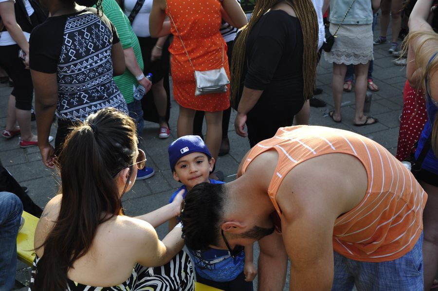 A young and happy male child glances up at the camera while in the middle of the dancefloor. His male and female parents are bending over to adjust his clothing and assist with his needs.