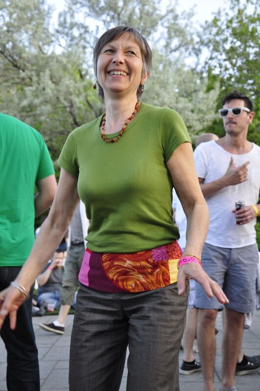 An older white woman with short-cropped, white hair, dances while smiling. She is wearing a green shirt and black pants with tye-dye patterns and a bead necklace.