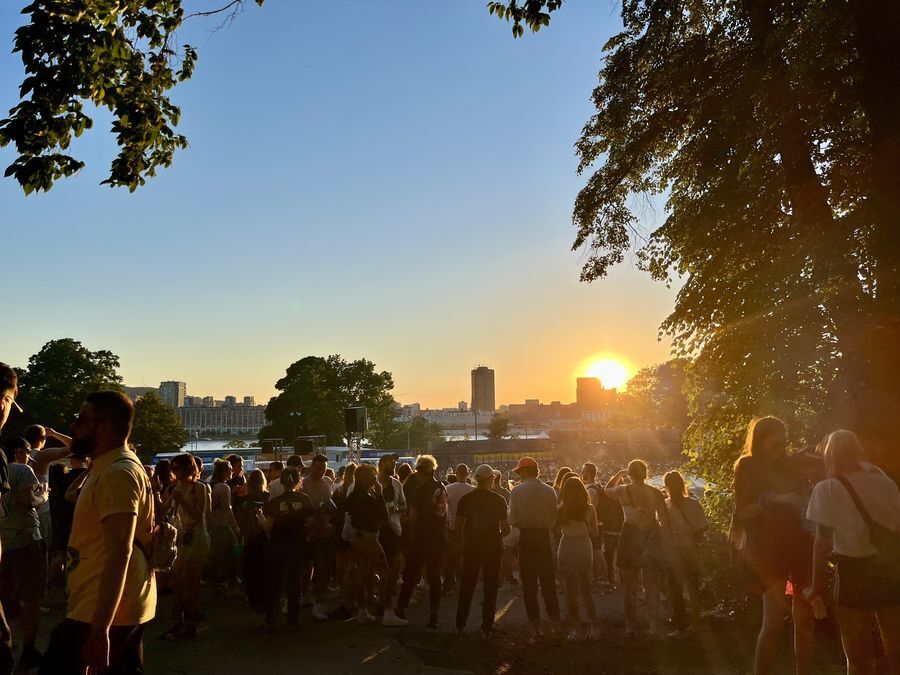 A crowd of people faces the sun, which is setting behind Montréal’s downtown skyline. The people are surrounded by trees.