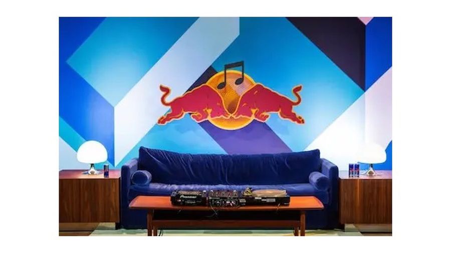 A stage is set for an informal lecture. In the foreground is a DJ setup on a coffee table and a purple sofa. A large logo for Red Bull is positioned in the background, while cans of Red Bull sit on tables either side of the sofa